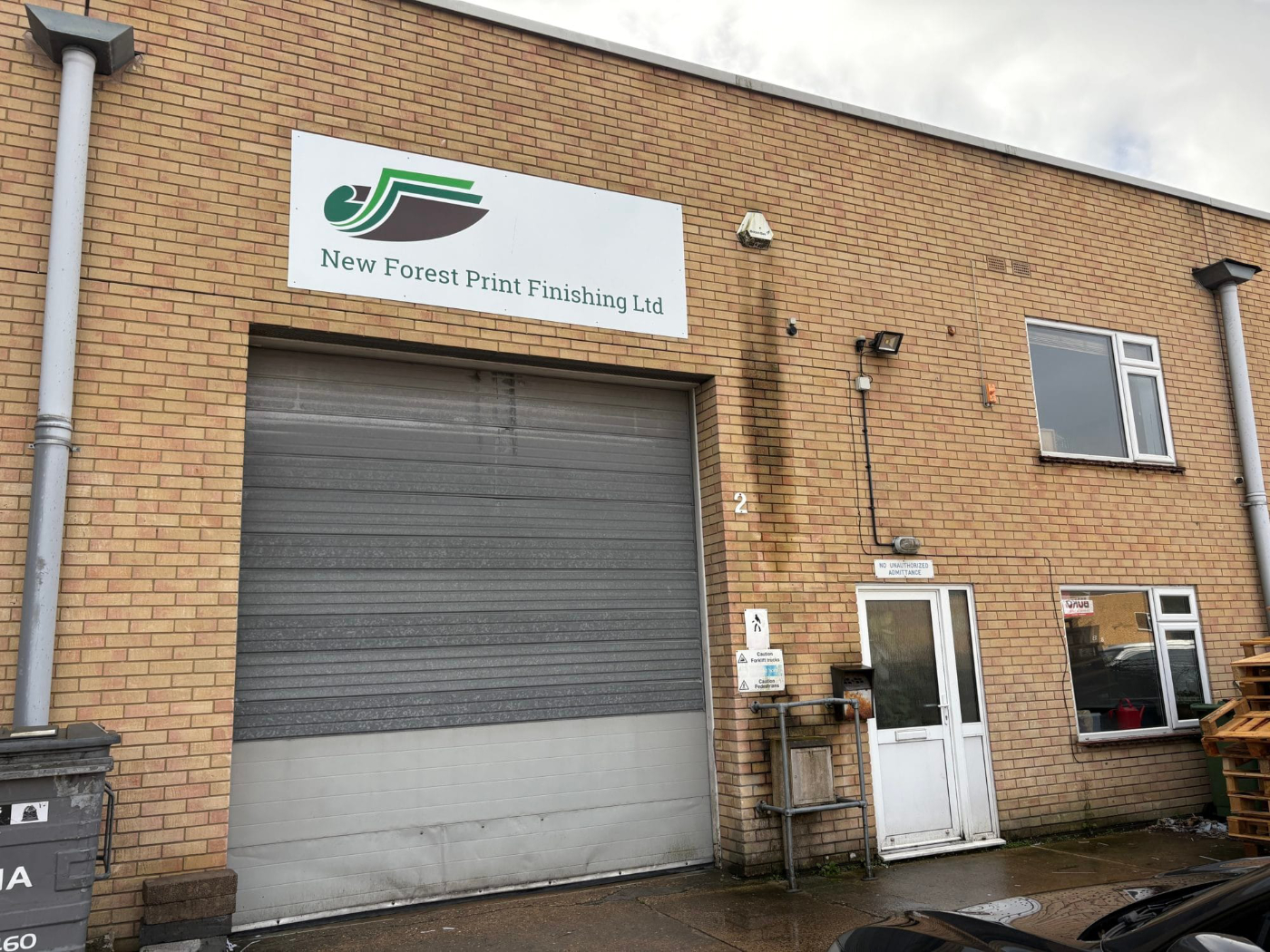 Exterior view of a brick industrial unit with roller shutter door and signage for New Forest Print Finishing Ltd above the entrance