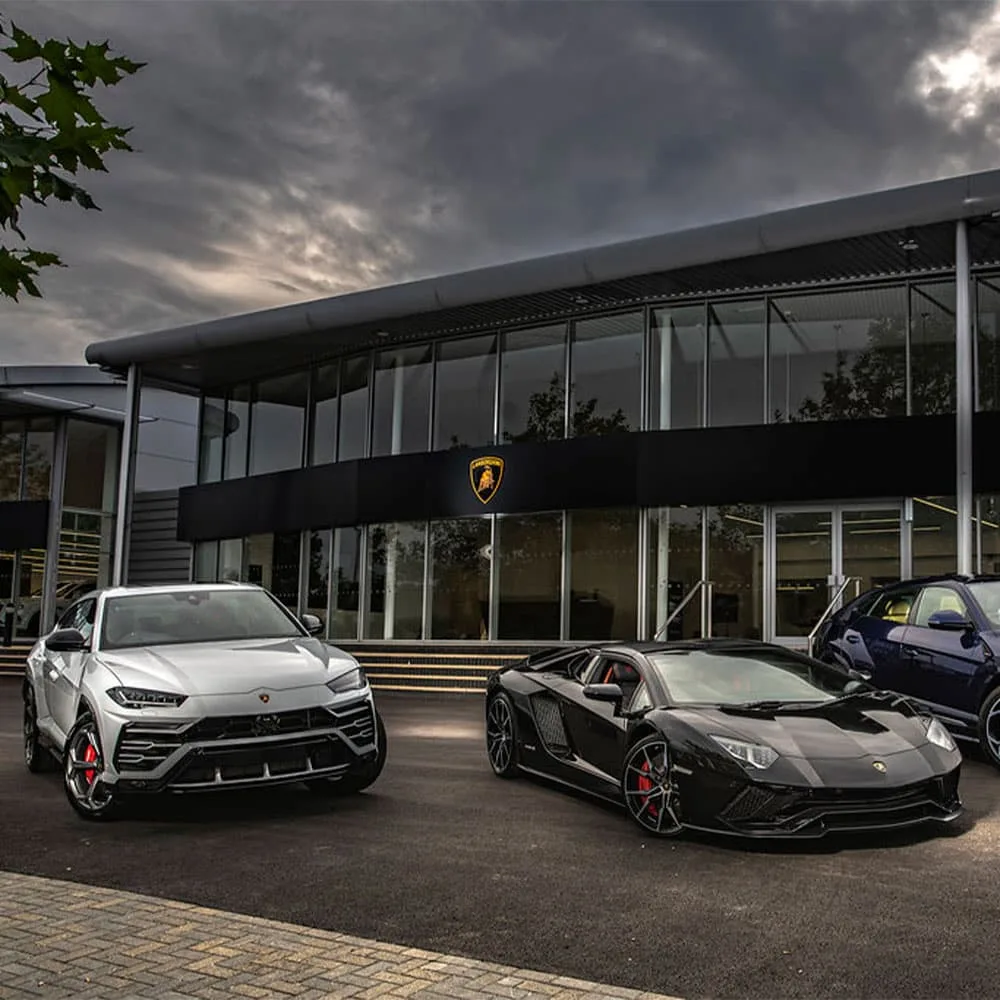 Exterior view of a Lamborghini dealership with luxury sports cars displayed on the forecourt in front of a modern showroom building.