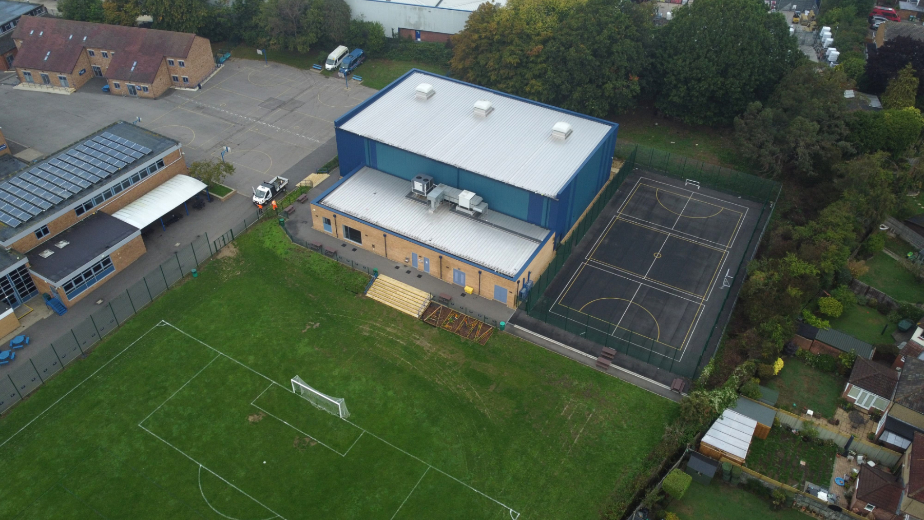 Aerial drone view of a school sports facility showing a sports hall building, outdoor multi-use games area, football pitch and surrounding school grounds.