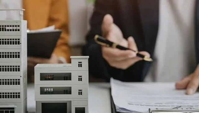 Two people sat at table with model of building on table and hand pointing at clip board.