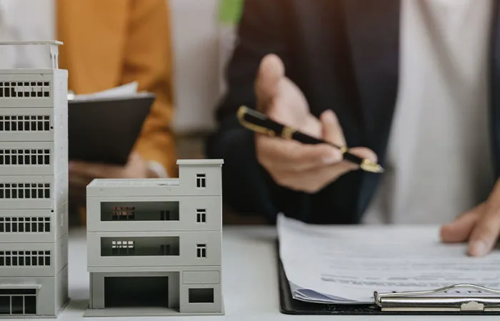 Two people sat at table with model of building on table and hand pointing at clip board.