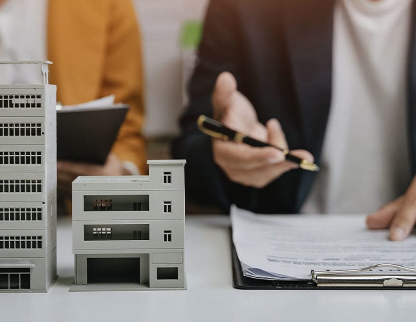 Two people sat at table with model of building on table and hand pointing at clip board.