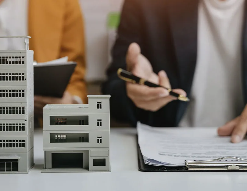 Two people sat at table with model of building on table and hand pointing at clip board.