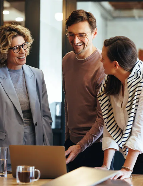 Three professionals collaborating around a laptop in an office meeting, discussing work in a modern workplace setting.