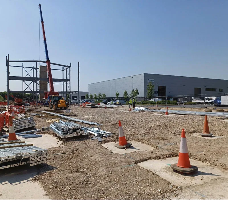 Early-stage commercial construction site with crane lifting steel framework, ground preparation works and materials laid out prior to building development