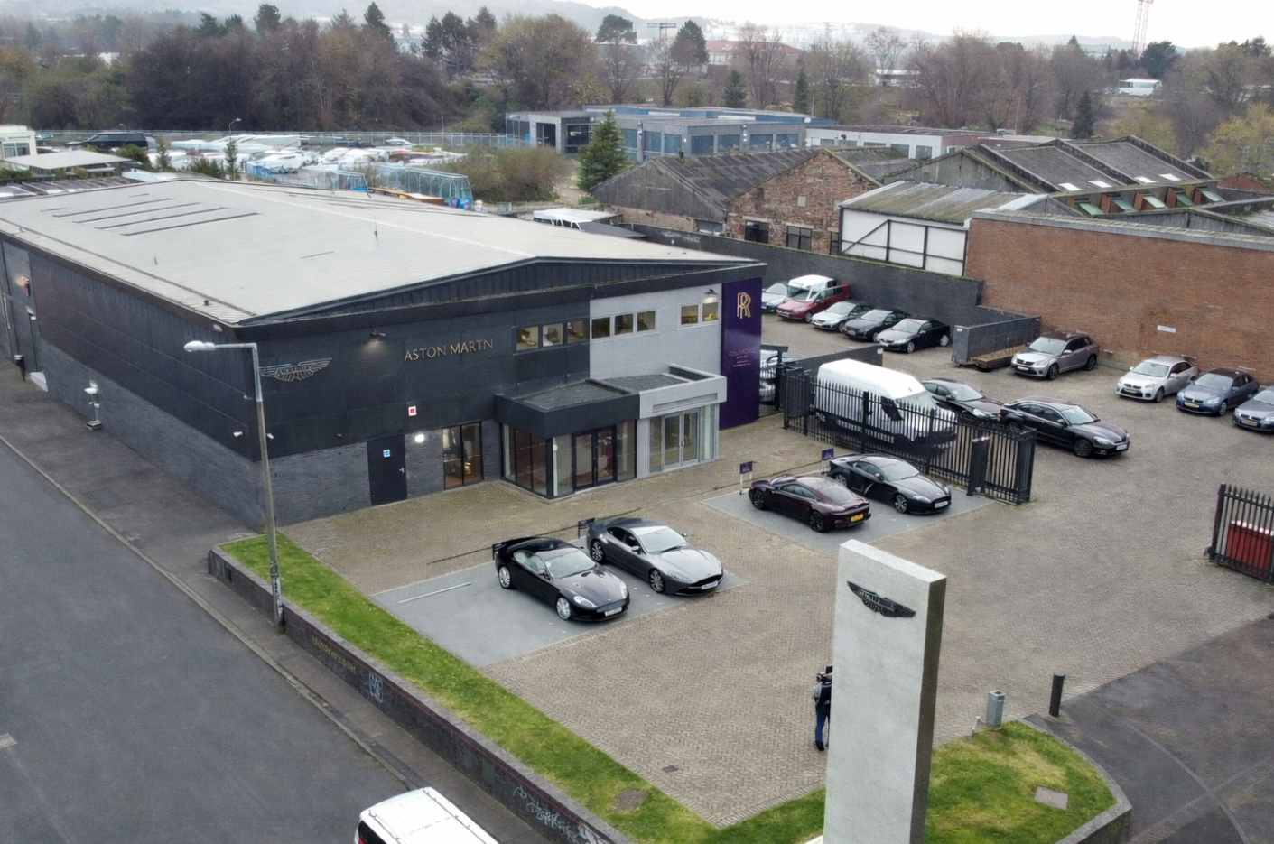 Aerial view of an Aston Martin car dealership with showroom building, forecourt parking and surrounding commercial premises.