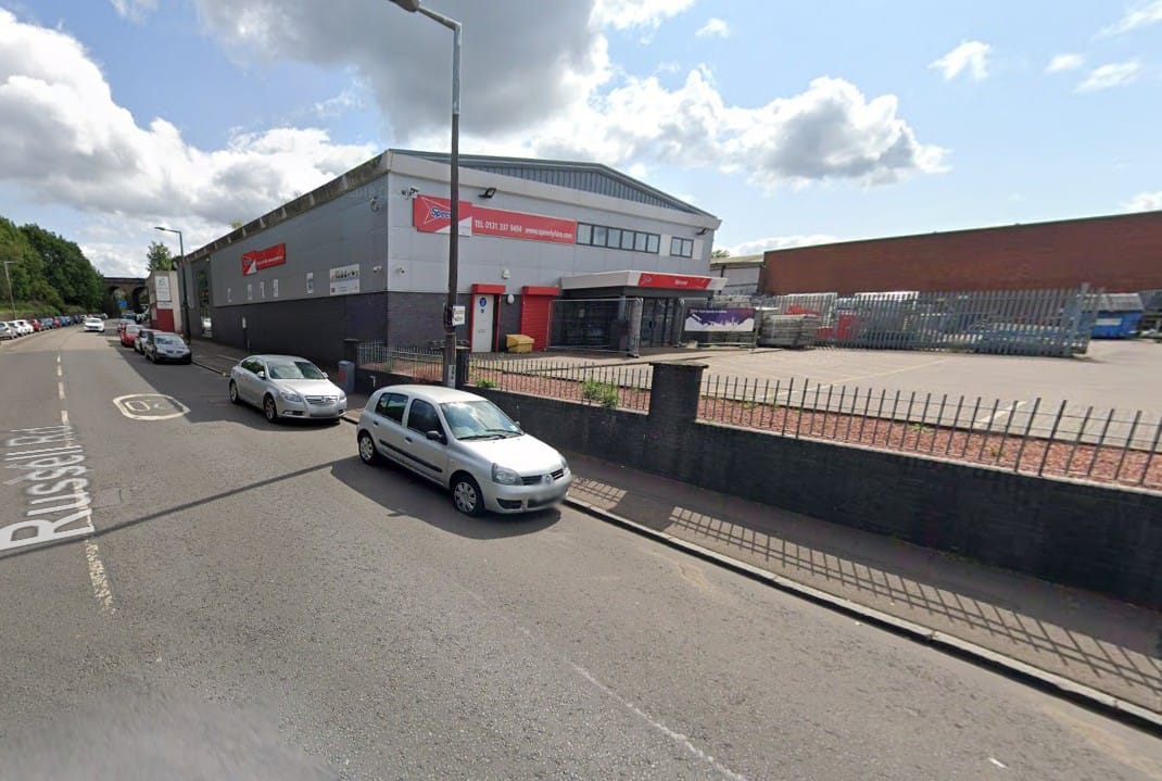 Street view of a commercial warehouse and trade counter unit with parking area and vehicles along an urban roadside.
